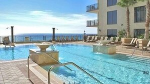 Uma área de piscina com vista clara para o oceano, cadeiras de lounge e um edifício com varandas.