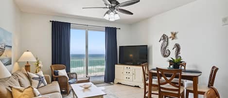 A well-lit room with a ceiling fan, a TV, a dining area with wooden chairs and a table, a sofa, and a large window with curtains.