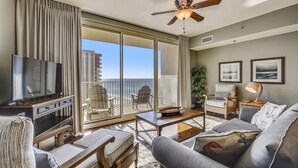 A spacious living room with a large TV, comfortable seating, a ceiling fan, and a view of the ocean.