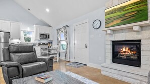A modern living room with a fireplace, leather sofa, dining area, kitchen, and a large flat-screen TV. - Appian Way Maltese Cottage 1 Bedroom Cabin by RedAwning (Fredericksburg)