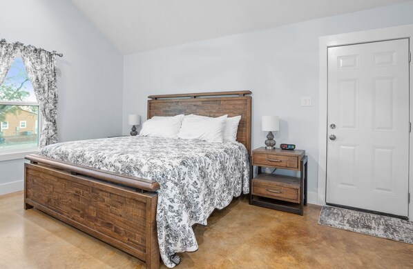Un dormitorio con una cama de madera, una mesita de noche, un cajón, una puerta y una ventana con cortinas.
