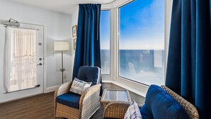 A room with a large window, a wicker chair, a small table, and a lamp. - Emerald Beach 1830 - 2 Bedroom/2 Bath+Bunks 2 Condo by RedAwning (Panama City Beach)