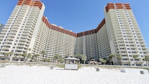Um hotel alto com várias varandas, uma área de praia com palmeiras e um pequeno coreto.