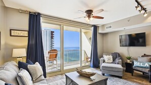 Ampla sala de estar com uma grande porta de vidro deslizante que dá para uma varanda com vista para o mar. Há um sofá confortável, uma mesa de centro e uma TV montada na parede.