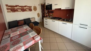 Une cuisine compacte avec des meubles blancs, une cuisinière à gaz et un évier. On y trouve un coin repas avec une table recouverte d’une nappe à motifs et deux chaises en osier.