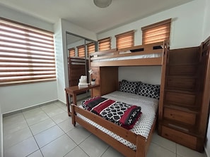 Ein Schlafraum mit Etagenbett aus Holz, einem kleinen Tisch und einem Fenster mit Jalousien.