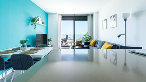 Ein modernes Wohnzimmer mit einem großen Glastisch, einer gelben Kissen ausgelegten Couch und Meerblick durch ein großes Fenster.