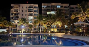Une vue nocturne d’un hôtel avec une piscine, des palmiers et un espace extérieur bien éclairé.