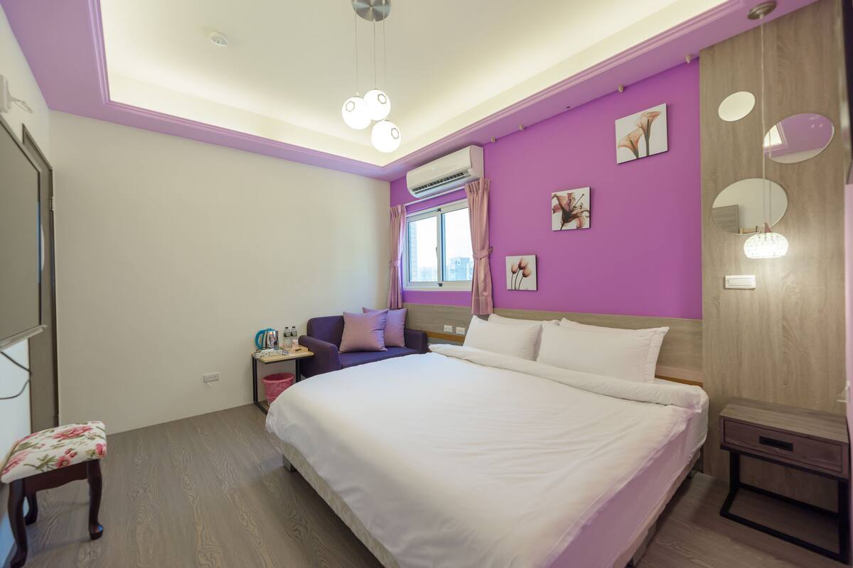 Quarto com uma cama grande, uma escrivaninha e uma cadeira. Há uma janela com cortinas, um ar condicionado e um lustre.