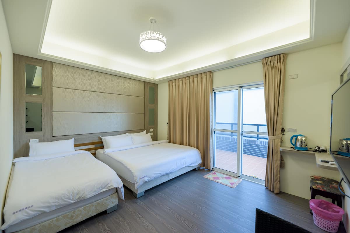 Quarto de hotel com duas camas, uma varanda com cortinas, uma escrivaninha com cadeira e uma pequena mesa com cesta.