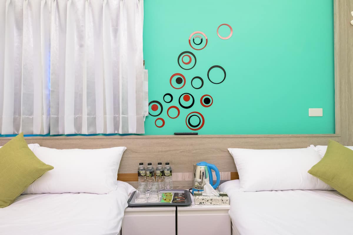 Quarto de hotel com duas camas, parede verde-água com padrões circulares, janela com cortinas brancas e mesa de cabeceira com produtos de higiene pessoal e caixa de lenços.