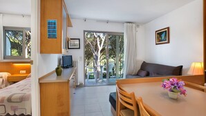 A compact room with a bed, a small desk with a computer, a dining table with chairs, a sofa, and a balcony with a view of trees. - Mas Vermey nº 6 - Apartment with shared swimming pool - Calella de Palafrugell (Calella de Palafrugell)