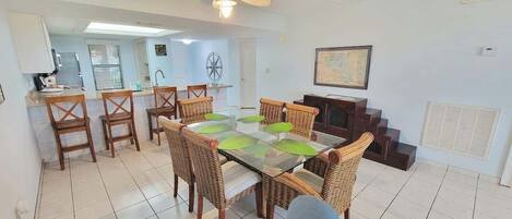 Un comedor con una mesa de vidrio rodeada de sillas de mimbre, una cocina con armarios blancos y una escalera que conduce a un nivel superior.