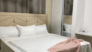 Un lit bien fait, avec une literie blanche et un plaid rose, une tête de lit beige et un grand miroir doté d’une porte coulissante.