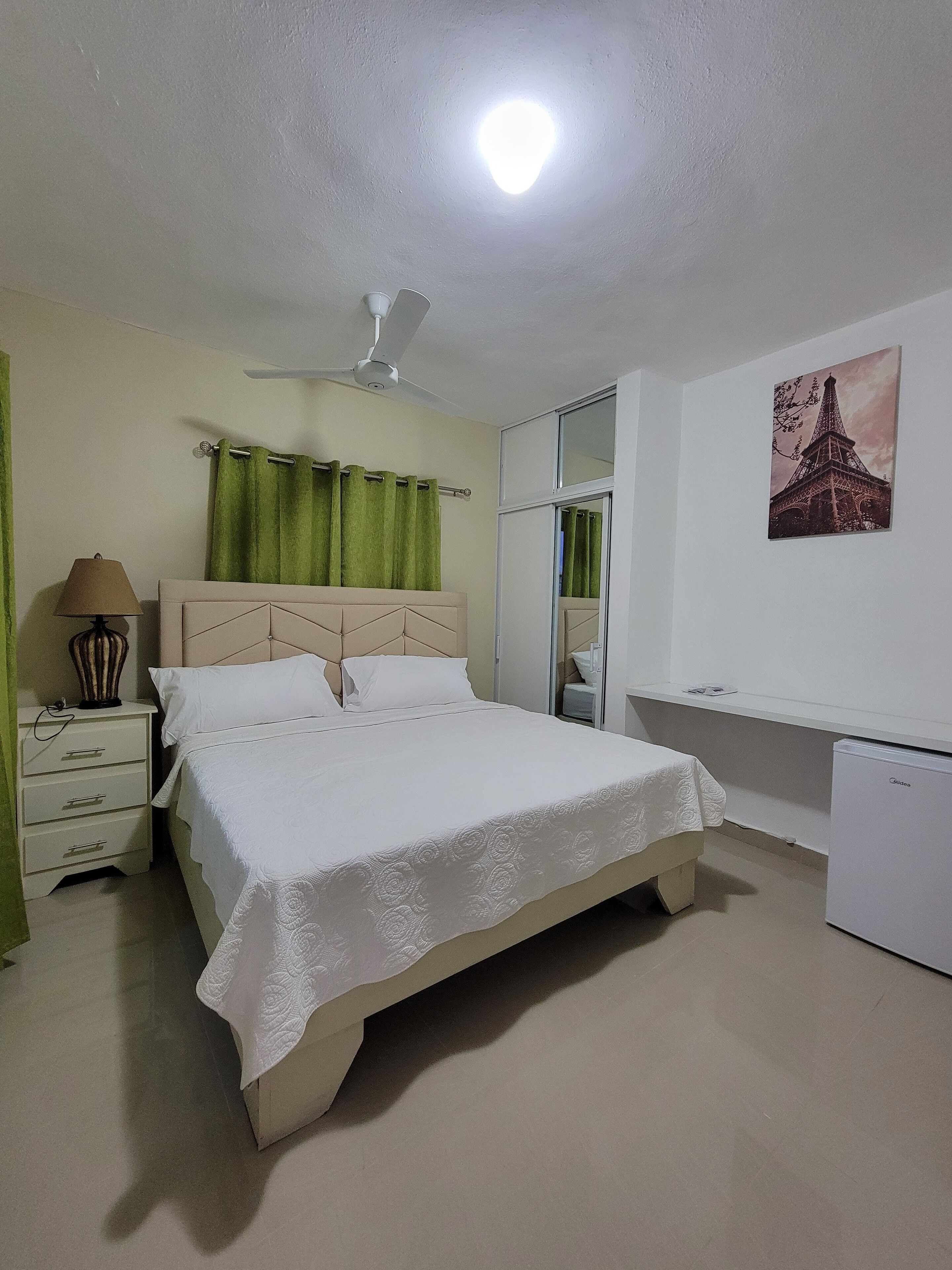 Une chambre à coucher comprenant un lit, une table de chevet, une lampe, un ventilateur de plafond, un rideau vert, une fenêtre et un réfrigérateur.