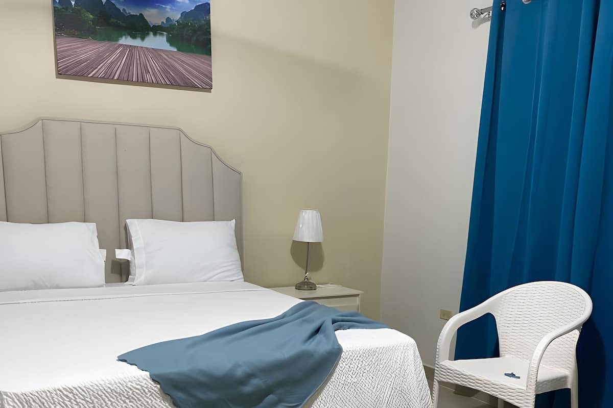 Un lit bien fait, avec du linge de lit blanc, un plaid bleu, une chaise blanche, une table de chevet avec une lampe et un tableau représentant un paysage accroché au mur.