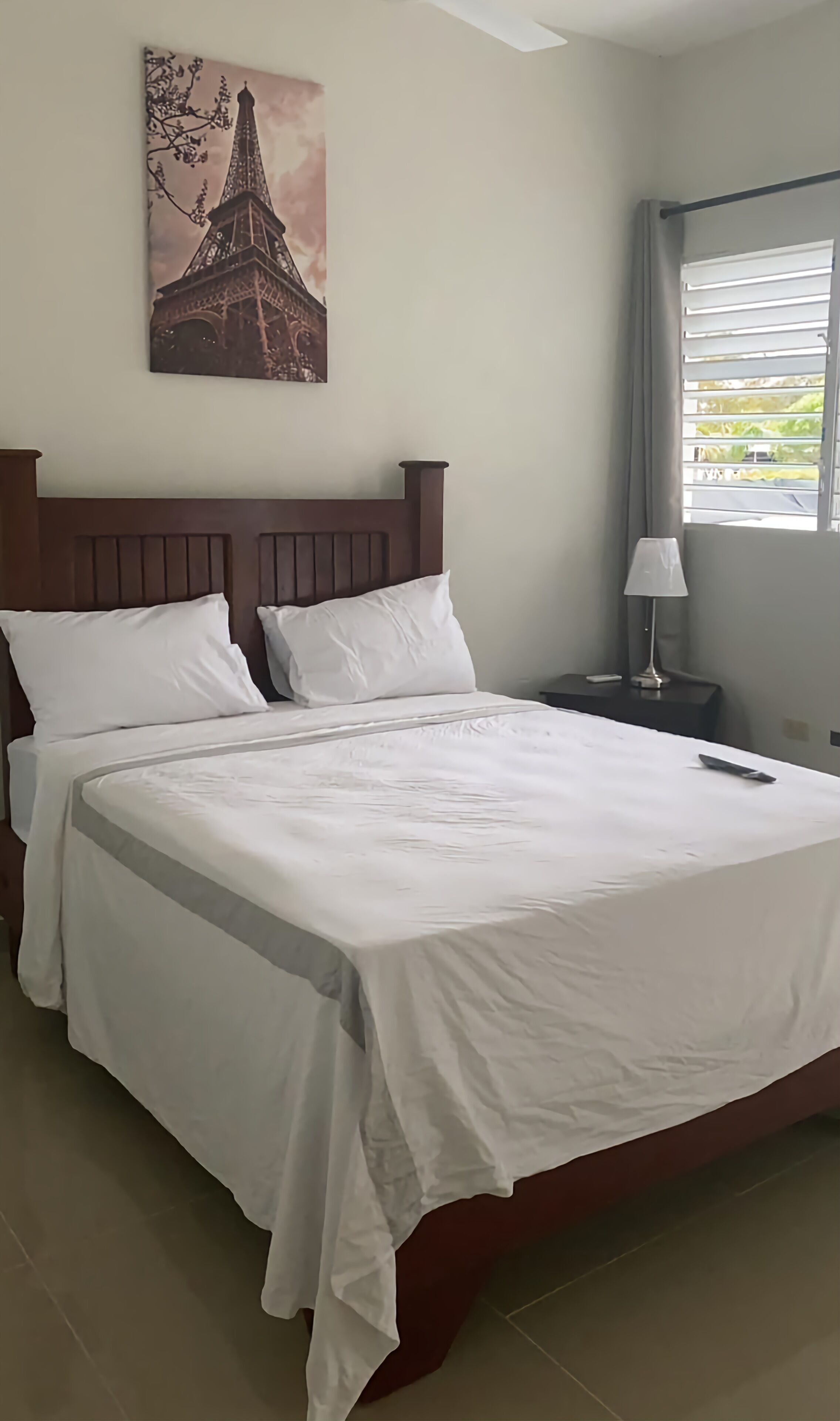A neatly made bed with white linens, a wooden headboard, and a painting of the Eiffel Tower on the wall.