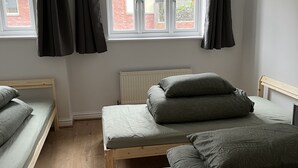 A bedroom with two single beds and a larger bed, a radiator, a window with curtains, and a view of a brick building outside.