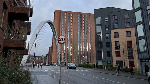 A modern urban street with a pedestrian bridge, a speed limit sign, and multi-story buildings.