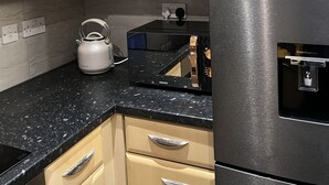 A kitchen with light wooden cabinets, a black countertop, a stainless steel refrigerator, and a kettle.