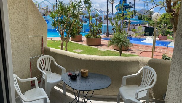 Een balkon met een tafel en stoelen, met uitzicht op een zwembadgedeelte met palmbomen en een blauwe glijbaan.