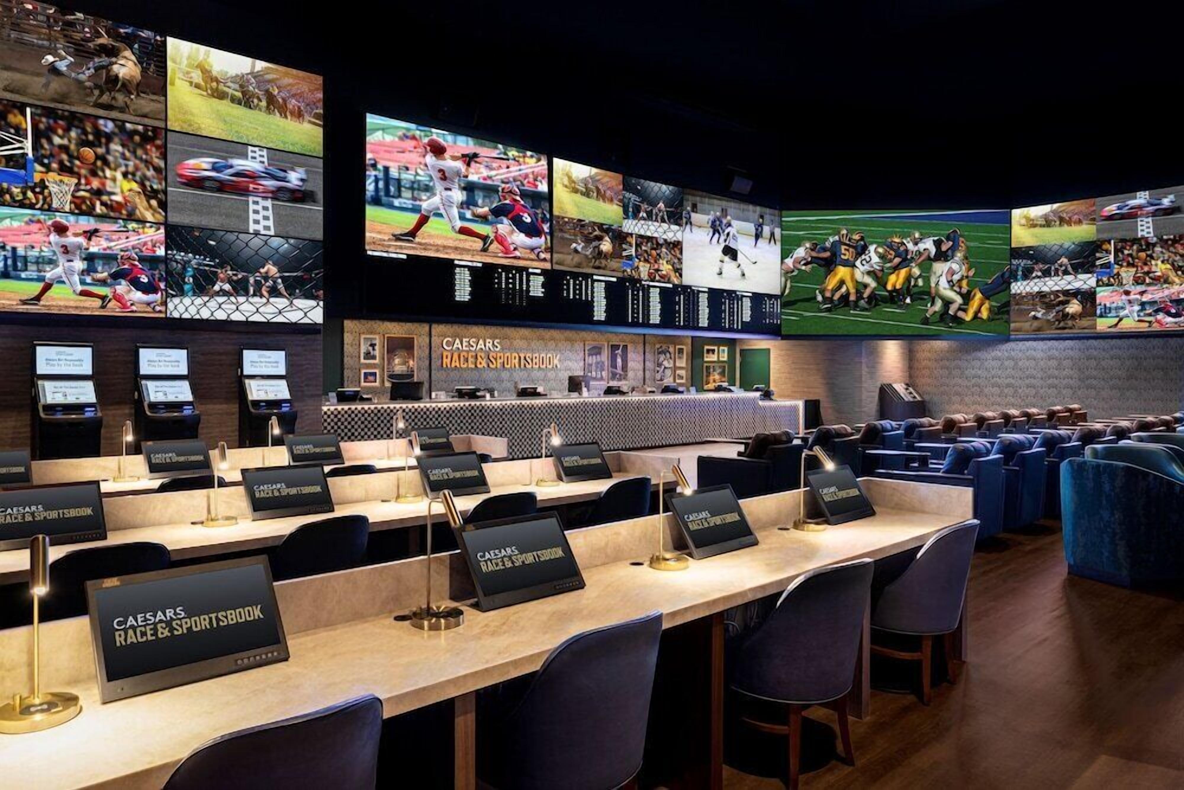 A sports-themed bar area with multiple screens displaying various sports events, a Caesars Race & Sportsbook sign, and a seating area with blue upholstered chairs.