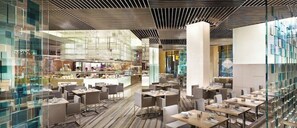 A modern restaurant with glass partitions, multiple dining areas, and a central kitchen visible through the partitions.
