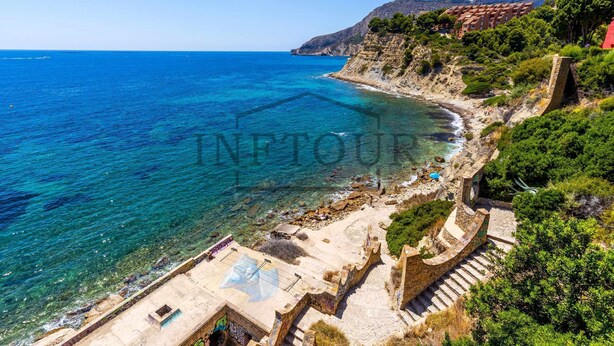 Un hotel frente al mar con fachada de piedra, una playa y montañas al fondo.