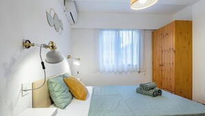 Un dormitorio con cama, almohadas, lámpara de escritorio, ventana con cortinas traslúcidas, armario de madera y unidad de aire acondicionado.