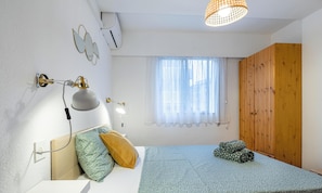A bedroom with a bed, pillows, a desk lamp, a window with sheer curtains, a wooden wardrobe, and an air conditioning unit.