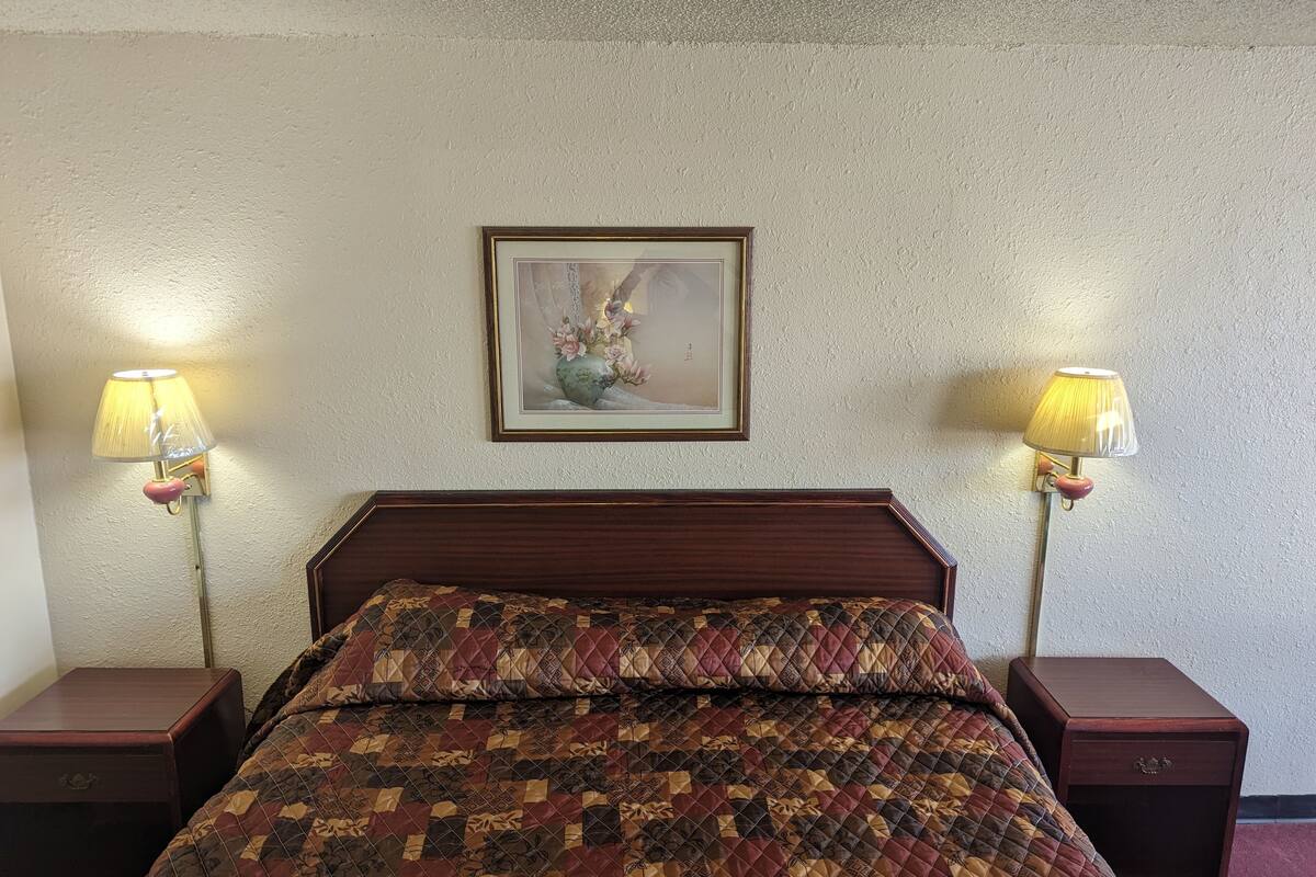 A bedroom with a wooden headboard, a quilted bedspread, two bedside tables with lamps, and a framed picture on the wall.