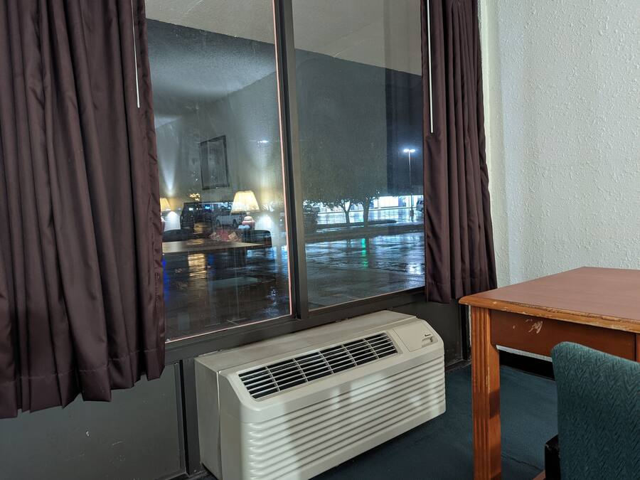 A room with a large window, a standing heater, a table, and a chair.