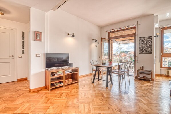 Een ruime woonkamer met een houten vloer, een flatscreen televisie, een eethoek met stoelen en een groot raam met uitzicht op de stad.