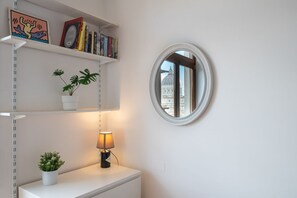 Een kamer met een ronde spiegel, een witte plank met boeken en planten, en een tafel met een lamp.
