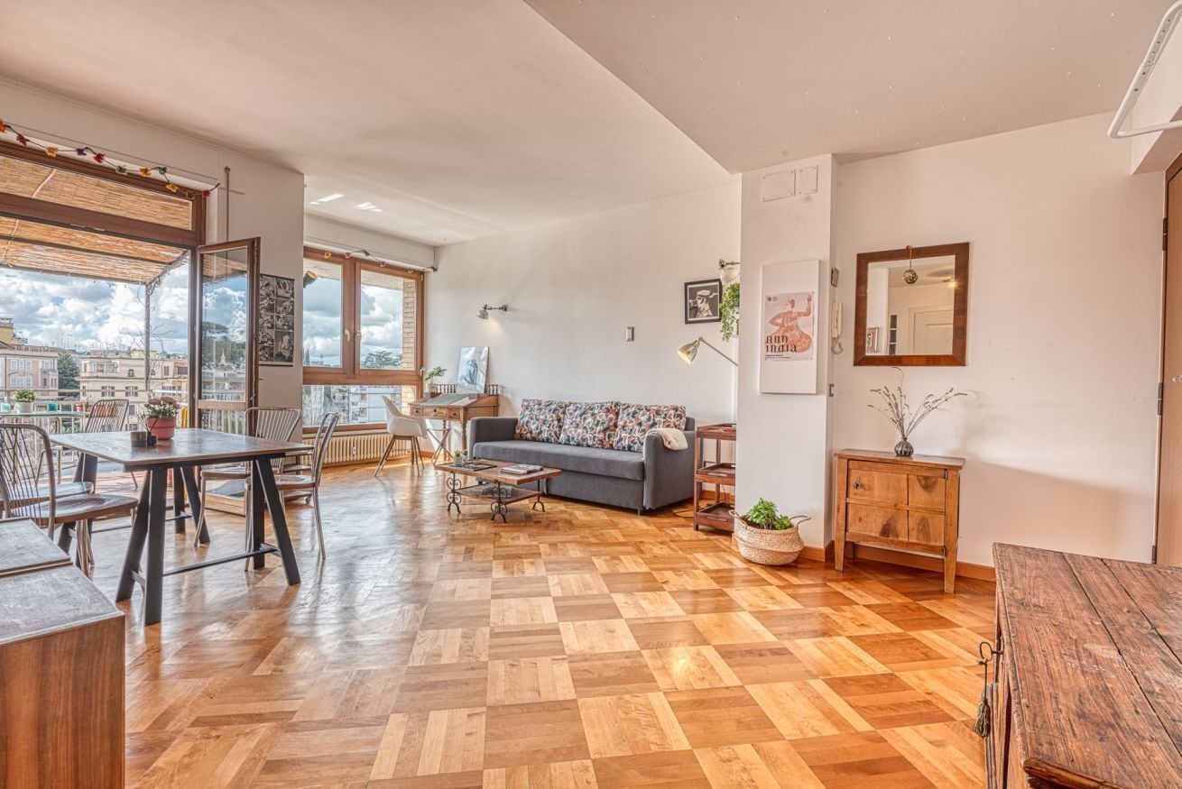 A spacious living area with a wooden parquet floor, a sofa, a dining table, and a view of the city through large windows.