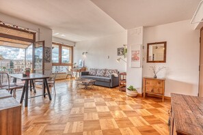 Een ruime woonkamer met een houten parketvloer, een bank, een eettafel en uitzicht op de stad door grote ramen.