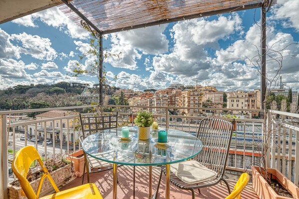 A balcony with a glass table, yellow chairs, and potted plants.