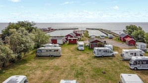 En campingplads med flere campingvogne parkeret på græsarealer, røde bygninger og udsigt til havet.