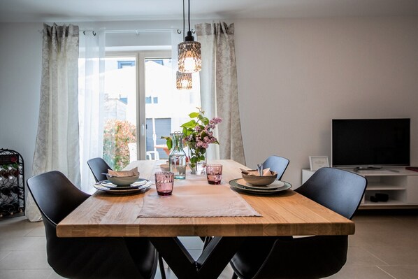 Een eethoek met een houten tafel, stoelen, een bloevas en een televisie op de achtergrond.
