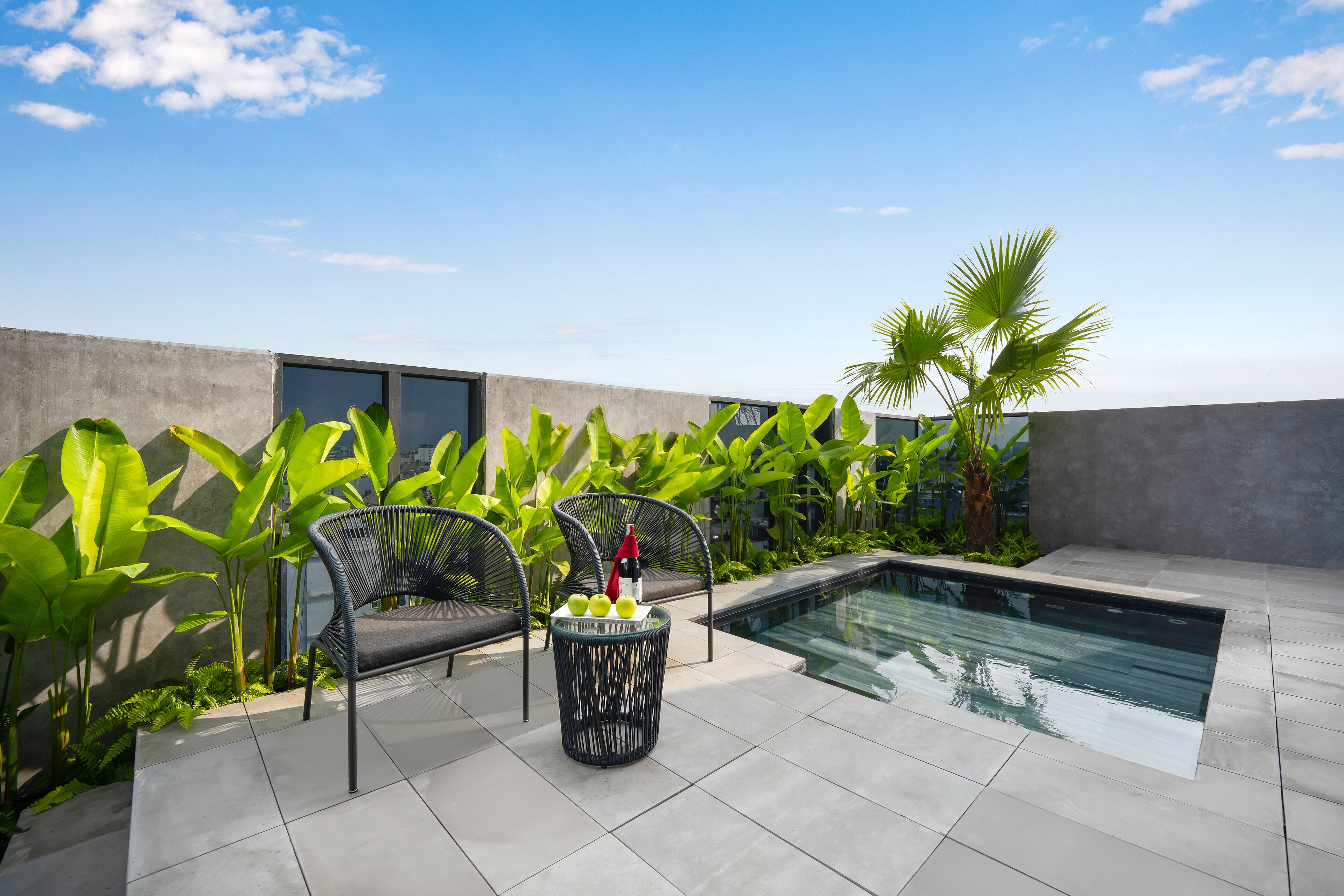 A rooftop pool area with two chairs, a small table with drinks, and lush greenery.