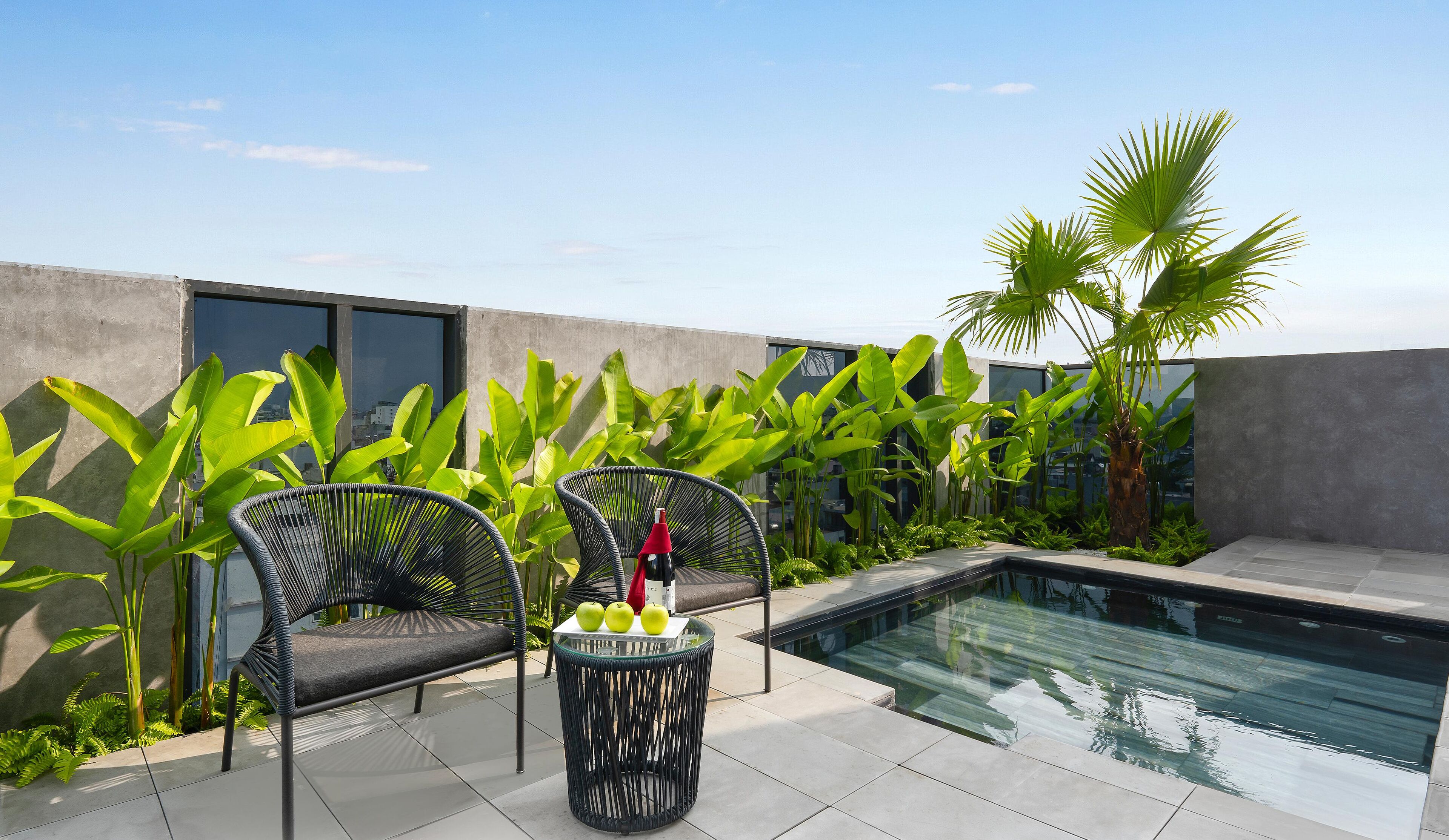 A rooftop pool area with two black wicker chairs, a small table with apples and a red bottle, surrounded by lush green plants and a modern building in the background.