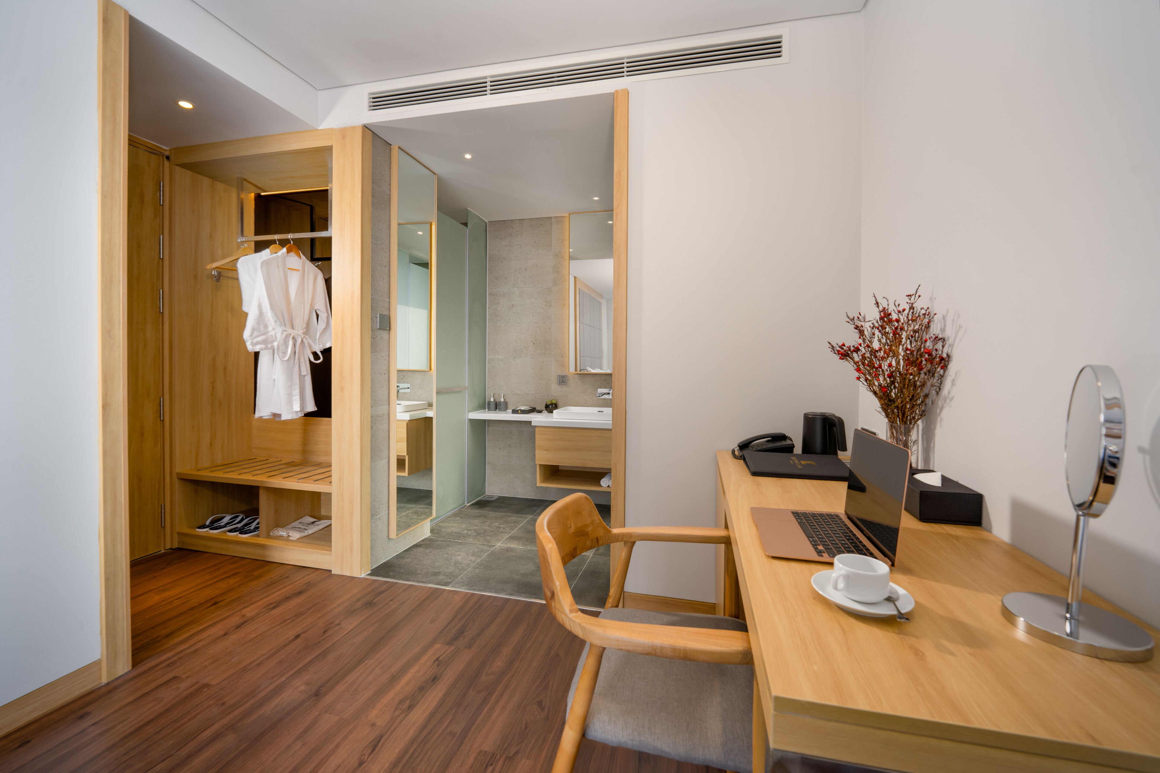 A modern hotel room with a wooden wardrobe, a desk with a laptop, a chair, and a small table with a cup and saucer.