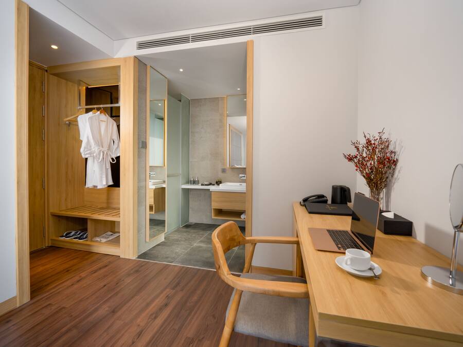 A modern hotel room with a wooden wardrobe, a desk with a laptop, a chair, and a small table with a cup and saucer.