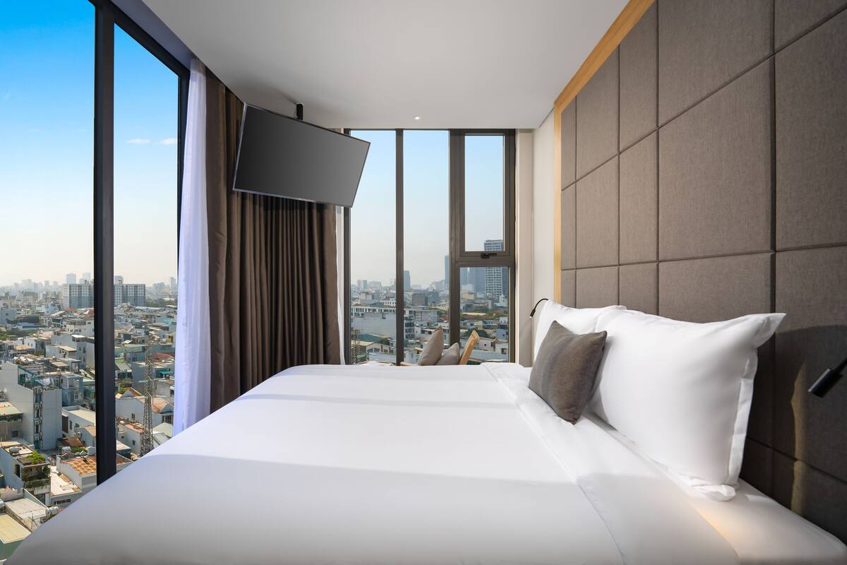 A hotel room with a large bed, a flat-screen TV, and a city view through large windows.