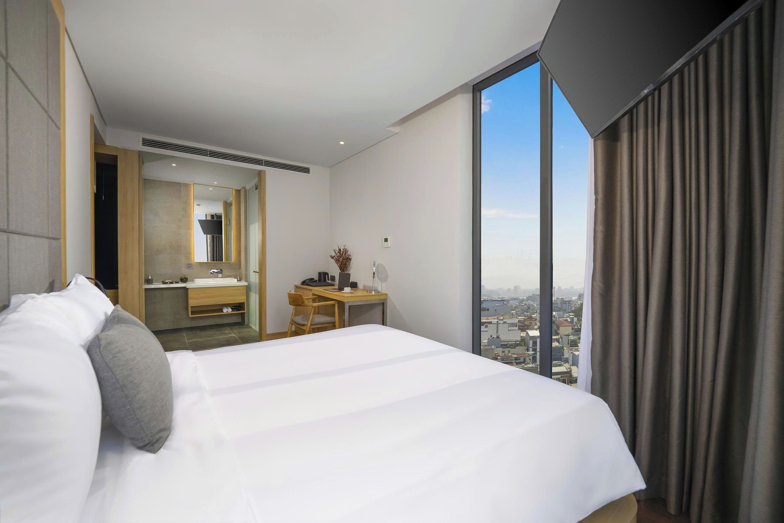 A hotel room with a bed, a desk, a bathroom, and a large window with a city view.