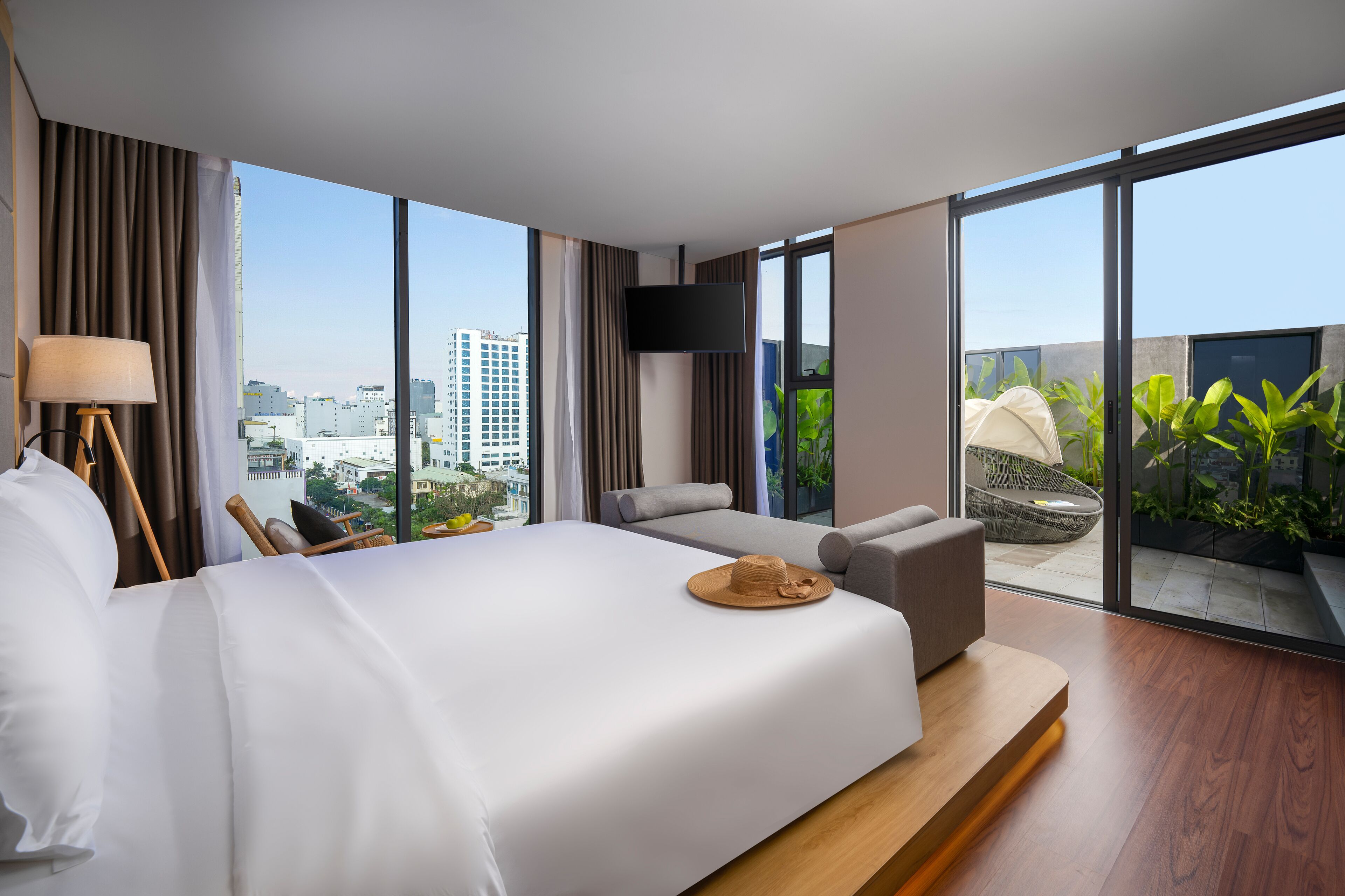 A modern hotel room with a large bed, a seating area, a TV, and a balcony with plants.