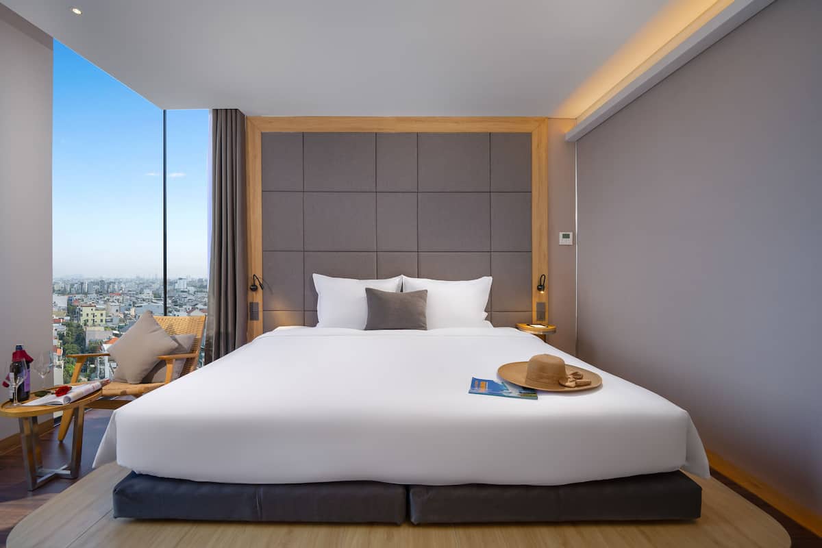 A modern hotel room with a large bed, a view of the city, and a balcony with a chair and table.