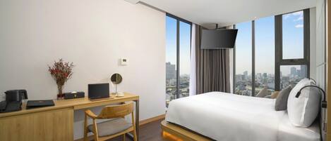 A hotel room with a bed, desk, chair, and a city view through large windows.