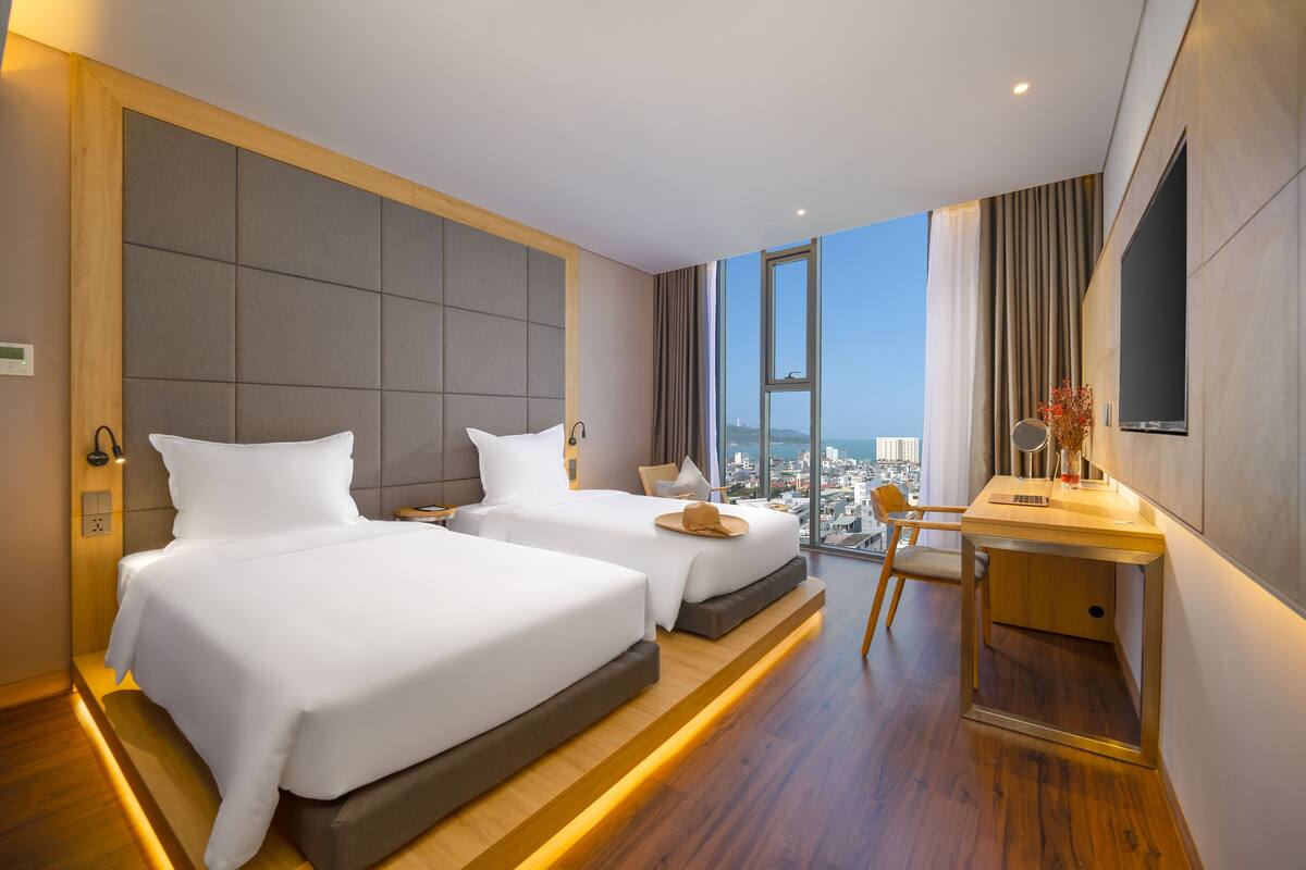A modern hotel room with two beds, a desk, and a large window offering a city view.