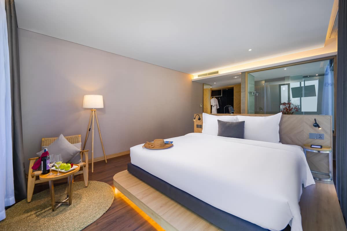 A modern hotel room with a large bed, a bedside table with a lamp, a chair with a small table, and a large mirror.